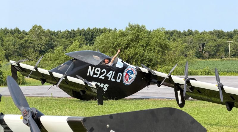 Pilot in a black biplane on a runway giving a thumbs-up, aircraft labeled N924LQ with an emergency services patch visible on the fuselage.