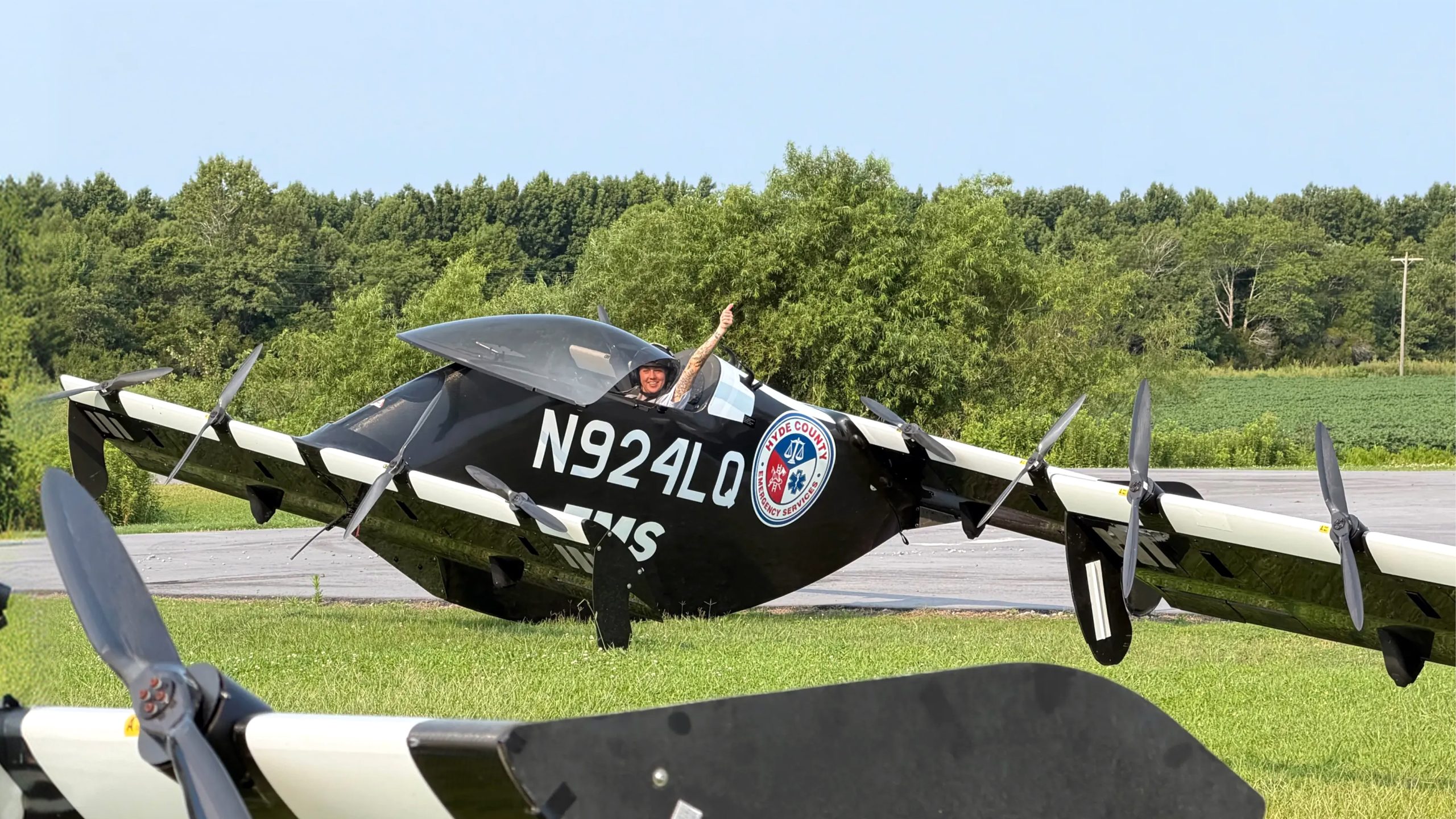 Pilot in a black biplane on a runway giving a thumbs-up, aircraft labeled N924LQ with an emergency services patch visible on the fuselage.