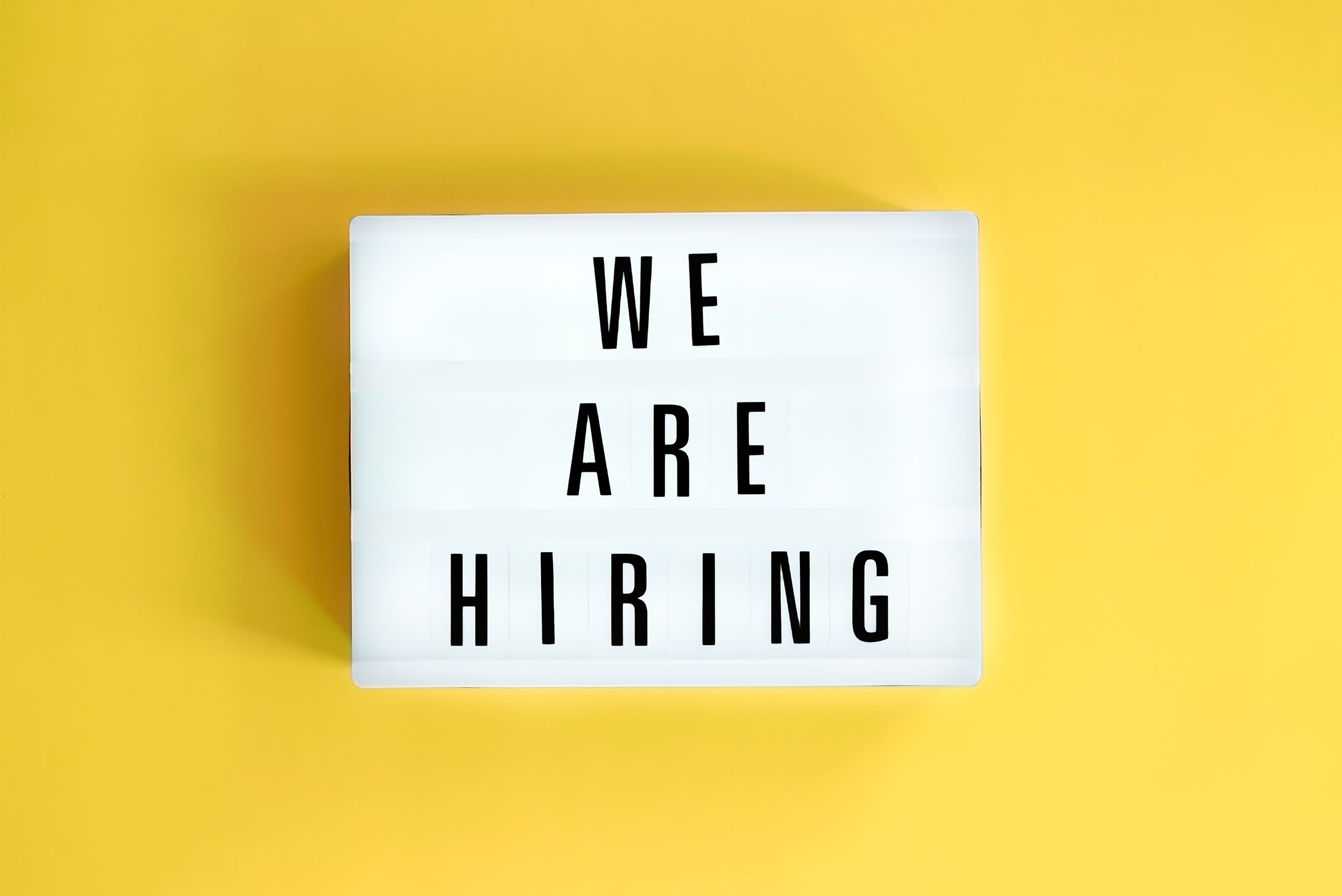 Lightbox sign on a yellow background reading 'WE ARE HIRING' in bold letters.