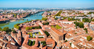 Aerial view of a city with a winding river, several bridges, and dense red-roofed buildings.
