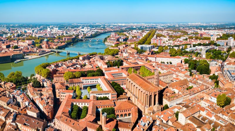 Aerial view of a city with a winding river, several bridges, and dense red-roofed buildings.