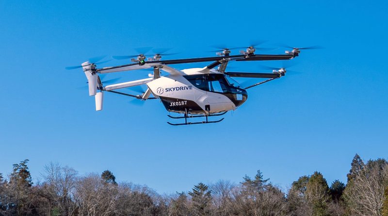SkyDrive six-rotor helicopter in flight above a treeline with a clear blue sky, front view showing its fuselage and landing skids.