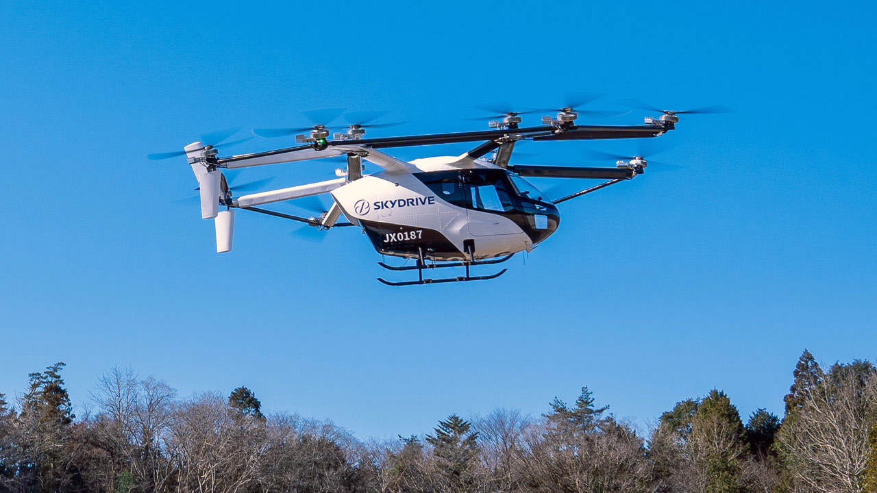 SkyDrive six-rotor helicopter in flight above a treeline with a clear blue sky, front view showing its fuselage and landing skids.
