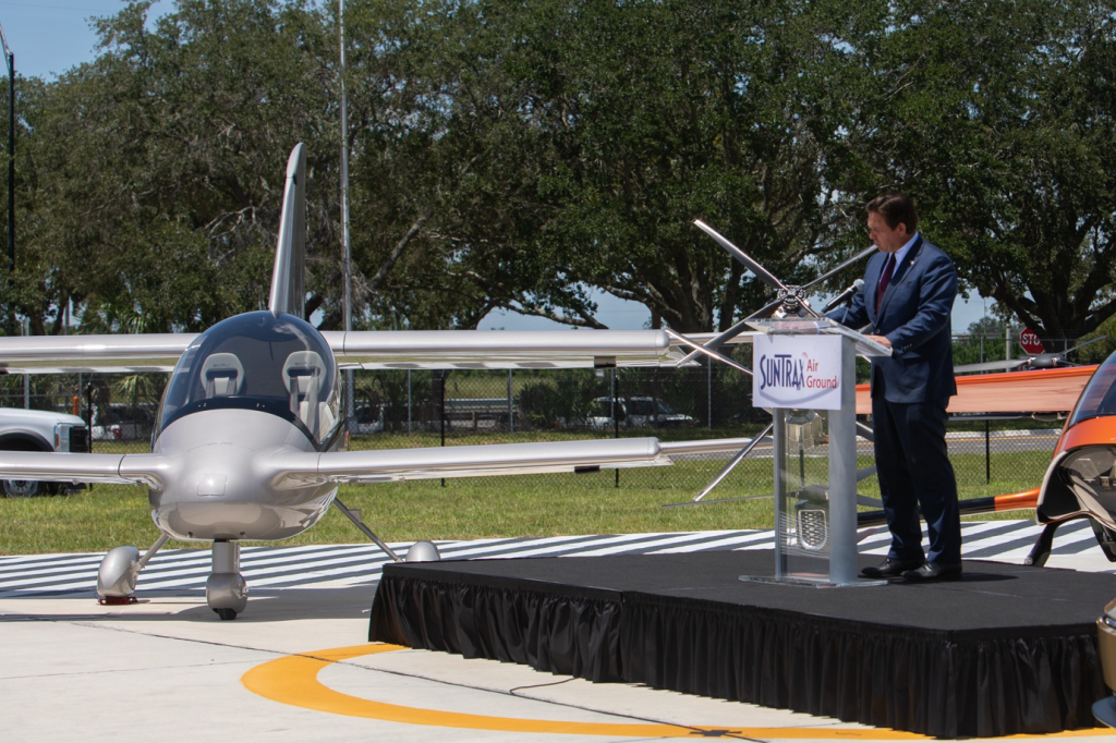 Man in a suit giving a speech at a podium on a black stage beside a small silver aircraft on a sunny day at an air show or dedication event, with trees in the background and a banner on the lectern.