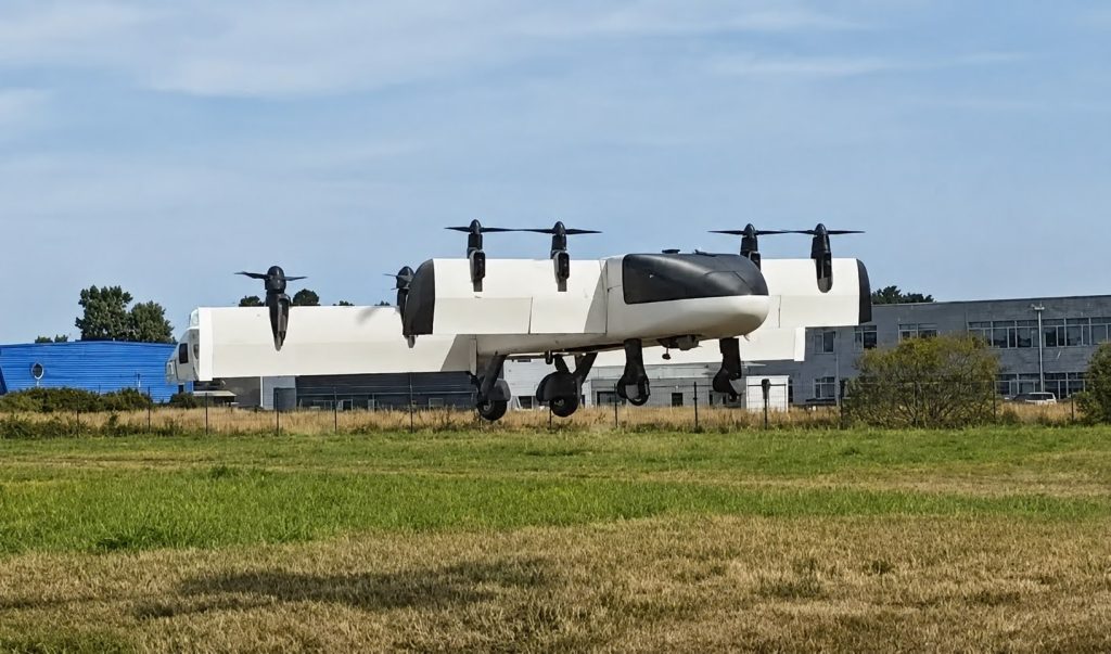 upscaled - eVTOL Insights White tilt-rotor aircraft with four propellers raised on landing gear, resting on a grassy airfield with industrial buildings in the background.