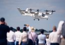 Tiltrotor Osprey aircraft flying low over spectators at an air show, crowd capturing the moment with phones and cameras.