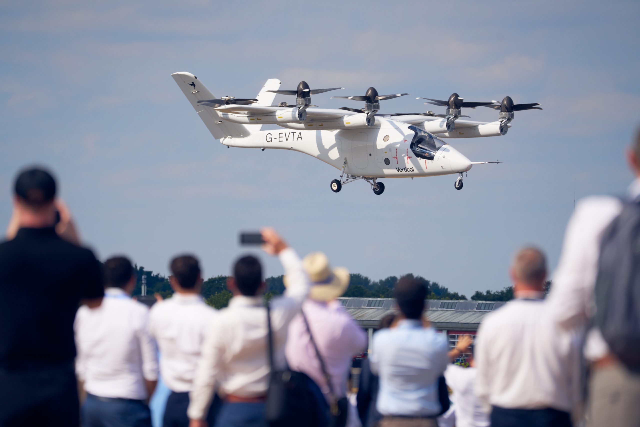 Tiltrotor Osprey aircraft flying low over spectators at an air show, crowd capturing the moment with phones and cameras.